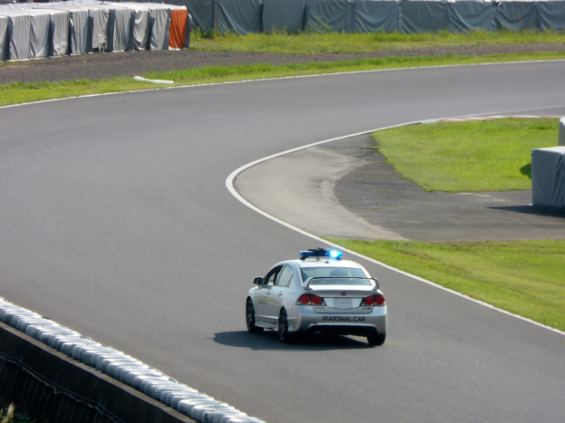 Polizeiauto fährt eine Rennstrecke entlang, umgeben von Gras auf beiden Seiten, Menschen und Zelte sind im Hintergrund zu sehen.