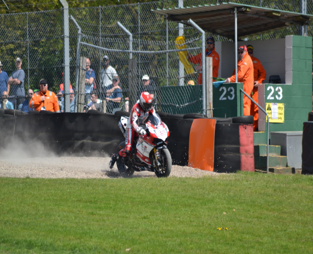 Ein Motorradfahrer in Helm und Handschuhen fährt über ein grasbewachsenes Feld, mit Zuschauern in Mützen und Schutzbrillen, einem eingezäunten Bereich, Treppen mit Geländern und einem nummerierten Schuppen im Hintergrund.