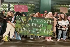 Eine Gruppe von Menschen hält ein Transparent mit der Aufschrift "Kein Feminismus ohne Trans-Frauen" und Schilder in einer Öffentlichen Straße mit einer Wand und einem Rollladen im Hintergrund.