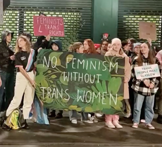 Eine Gruppe von Menschen hält ein Transparent mit der Aufschrift "Kein Feminismus ohne Trans-Frauen" und Schilder in einer Öffentlichen Straße mit einer Wand und einem Rollladen im Hintergrund.