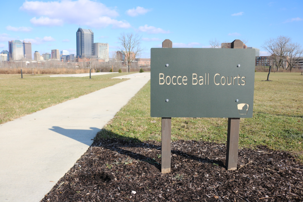 Ein Schild am Boden, das Boccia-Plätze in Indianapolis, Indiana, anzeigt, umgeben von Gras, Bäumen, einem Weg, Pfählen, einem Zaun, Gebäuden und einem bewölkten Himmel.