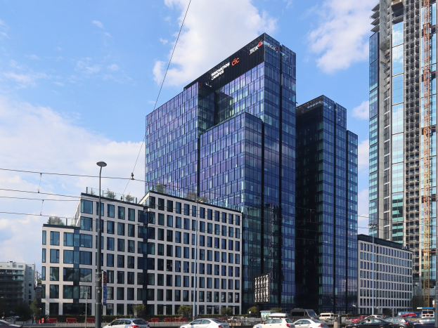 Großes Gebäude mit vielen Fenstern, die Deutsche Bank Hauptverwaltung in Frankfurt, Deutschland, umgeben von städtischen Elementen einschließlich Straßenlaternen, Fahrzeugen, Bäumen und einem bewölkten Himmel.