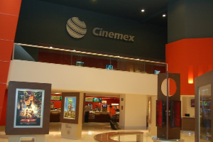Eine Gruppe von Menschen steht in einem Cinemex-Kino in einem Einkaufszentrum, mit einer Person, die sitzt, Tischen, Fotorahmen an der Wand, einem Schild, Deckenlampen und Geräten auf den Tischen.