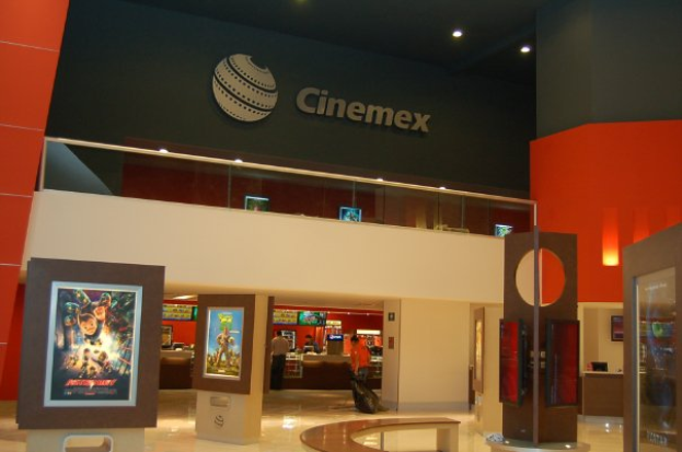 Eine Gruppe von Menschen steht in einem Cinemex-Kino in einem Einkaufszentrum, mit einer Person, die sitzt, Tischen, Fotorahmen an der Wand, einem Schild, Deckenlampen und Geräten auf den Tischen.