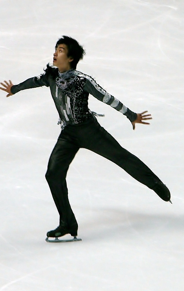 Männlicher Eiskunstläufer in schwarzer Kleidung gleitet über eine Eisbahn und führt eine Choreographie mit synchronen Arm- und Beinbewegungen aus.