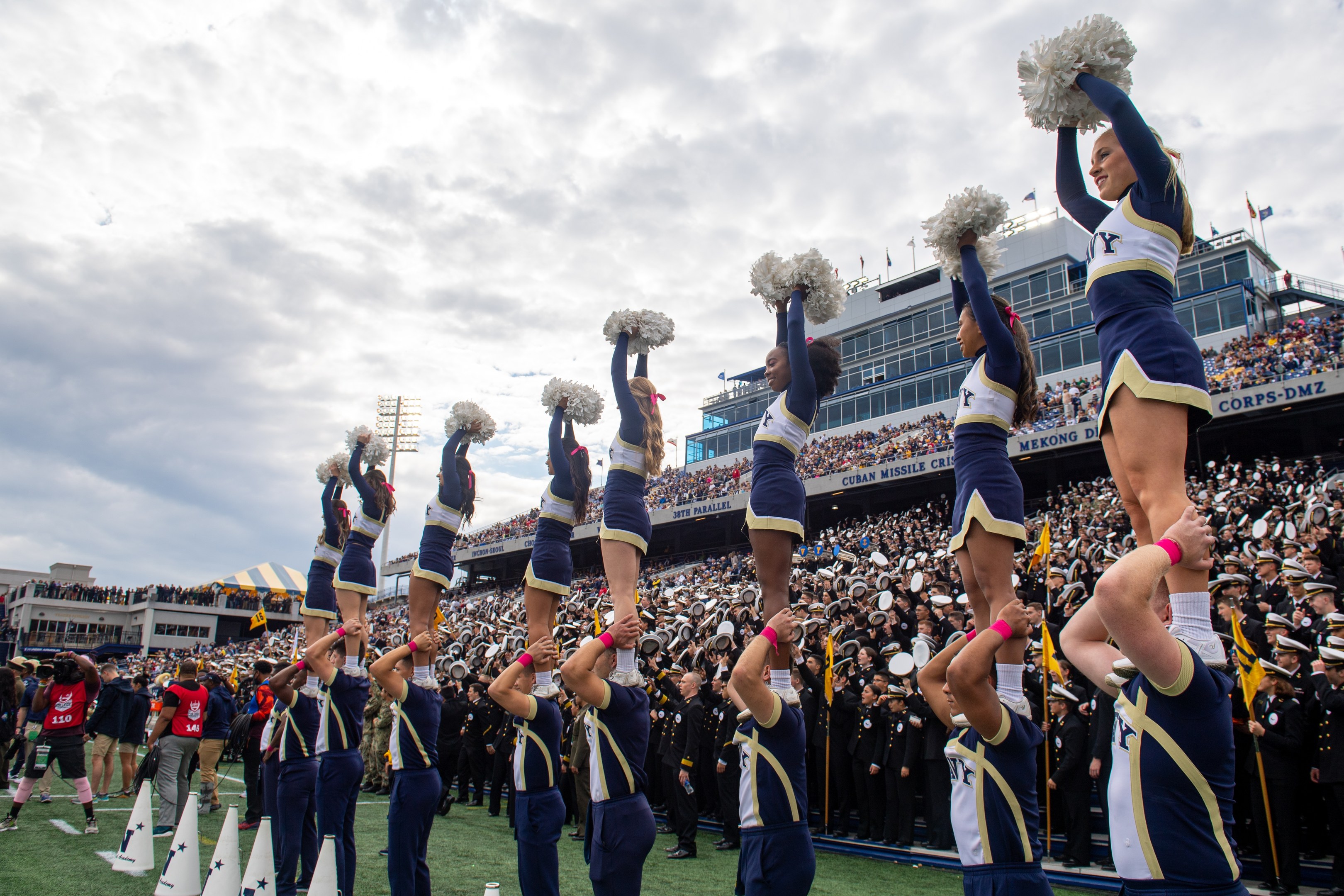 Eine Gruppe von Cheerleadern in blauen und weißen Uniformen führt einen Stunt auf einem Stadion-Event durch, während die Menge zuschaut und eine Person die Szene fotografiert.