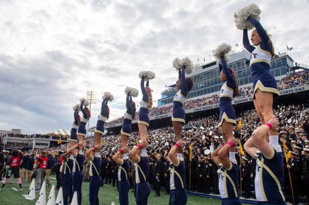 Eine Gruppe von Cheerleadern in blauen und weißen Uniformen führt einen Stunt auf einem Stadion-Event durch, während die Menge zuschaut und eine Person die Szene fotografiert.
