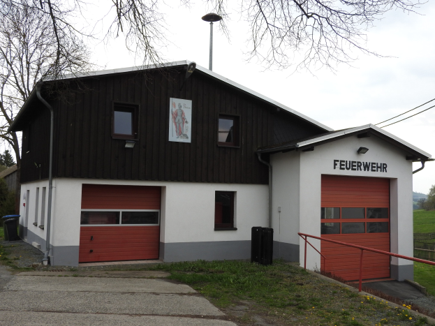 Feuerwache mit rotem und weißem Gebäude, Straße, Gras, gelben Blumen, schwarzen Objekten, Geländer, Pfahl, Drähte, Bäume und Himmel sichtbar.