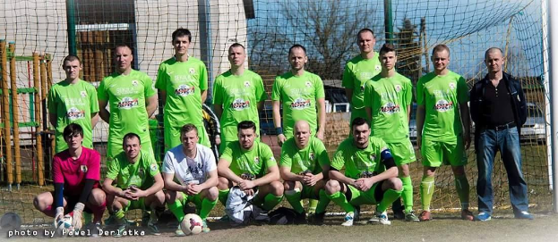 Gruppe von Männern in grünen T-Shirts auf einem Fußballfeld mit einem Ball vor ihnen, in der Nähe eines Torpfostens, mit Bäumen, Gebäuden und Fahrzeugen im Hintergrund.