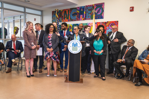 Eine Gruppe von Menschen steht vor einem Podium mit einem Mikrofon und einer Tafel mit der Aufschrift "Sancho Stadtratmann tritt dem Stadtrat bei", während andere auf Stühlen sitzen oder in der Nähe stehen; Flaggen, ein Feuerlöscher und Deckenleuchten sind im Hintergrund sichtbar.