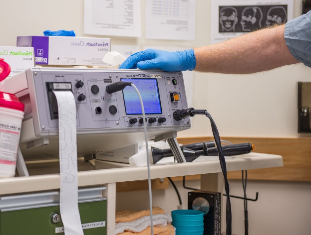 Eine Person in blauen Handschuhen arbeitet an einem medizinischen Gerät auf einem Tisch in einem unordentlichen Krankenhauszimmer mit Kartons, Tassen und Papieren an der Wand.