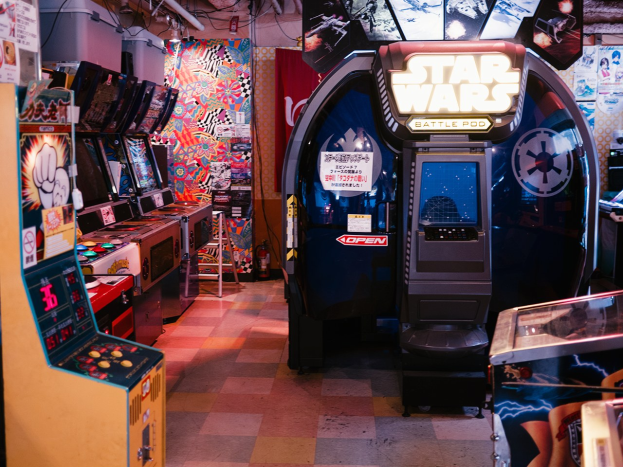 Ein hell erleuchteter Arcade-Raum mit mehreren Maschinen, darunter ein Star Wars Battle Pod, bunten Postern an den Wänden und verstreuten Gegenständen.
