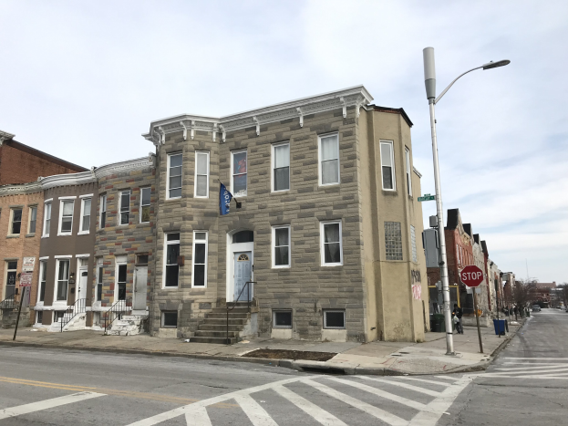Eine Reihe von Häusern mit Fenstern, Türen, Treppen mit Geländern, Laternen und Schildern an einer Straßenecke, mit Fahrzeugen auf der Straße, Bäumen im Hintergrund und einem bewölkten Himmel.