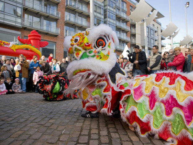 Ein lebendiges chinesisches Neujahrsfest in Amsterdam mit einer Löwen-Tanzvorstellung vor einer Zuschauermenge, einige halten Kameras, vor dem Hintergrund von Gebäuden, Laternenmasten und einem klaren blauen Himmel.