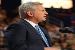 Ein Mann im Anzug und mit Krawatte steht an einem Podium mit zwei Mikrofonen und sieht ernst aus, mit einem unscharfen Hintergrund.