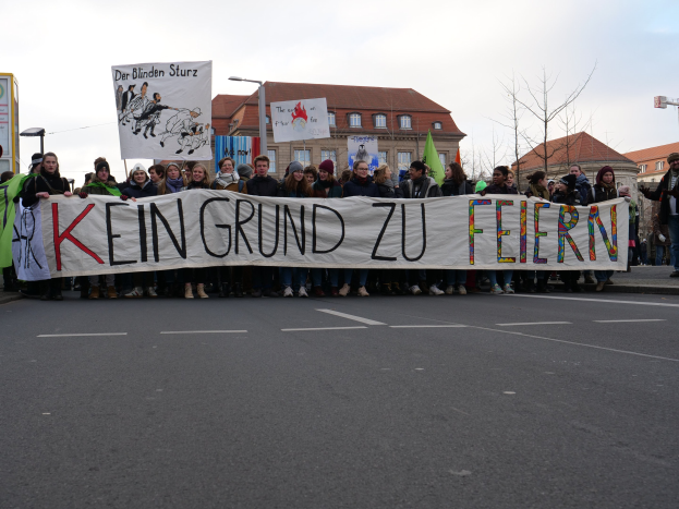 Demonstranten mit einem Transparent mit der Aufschrift "Kein Grund zu Feiern" gegen deutsche Sparmaßnahmen, mit Gebäuden, Laternenmasten, Bäumen und einem klaren Himmel im Hintergrund.