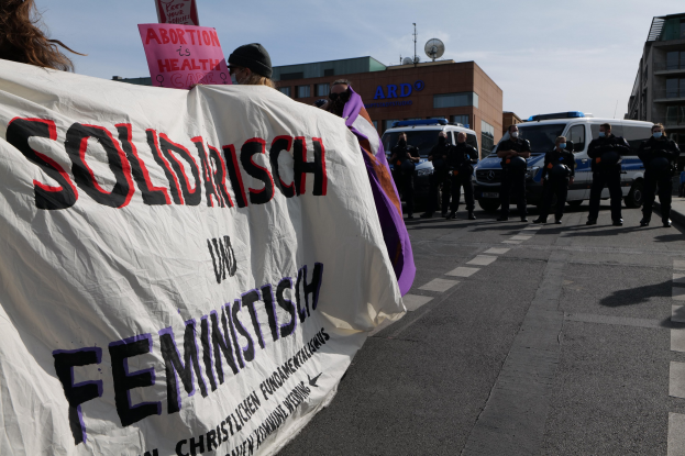 Gruppe von Menschen marschiert mit einem "Solidarität und Feminismus"-Schild die Straße entlang, mit parkenden Fahrzeugen, Gebäuden, einer Schüsselantenne und einem klaren blauen Himmel im Hintergrund.