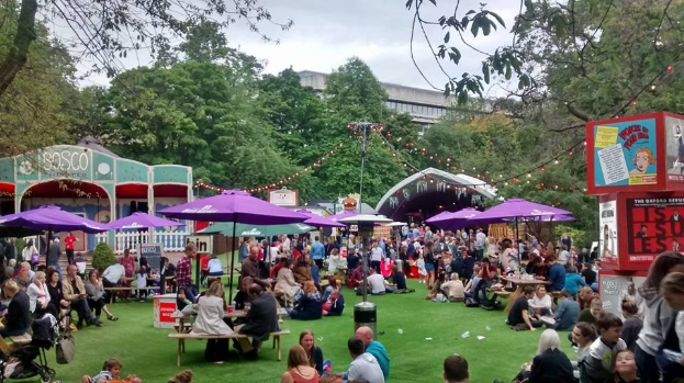 Große Gruppe von Menschen auf einem grünen Feld in einer parkähnlichen Umgebung mit Schirmen, Tischen und einer Tafel, die auf ein Biergarten in London hinweist.