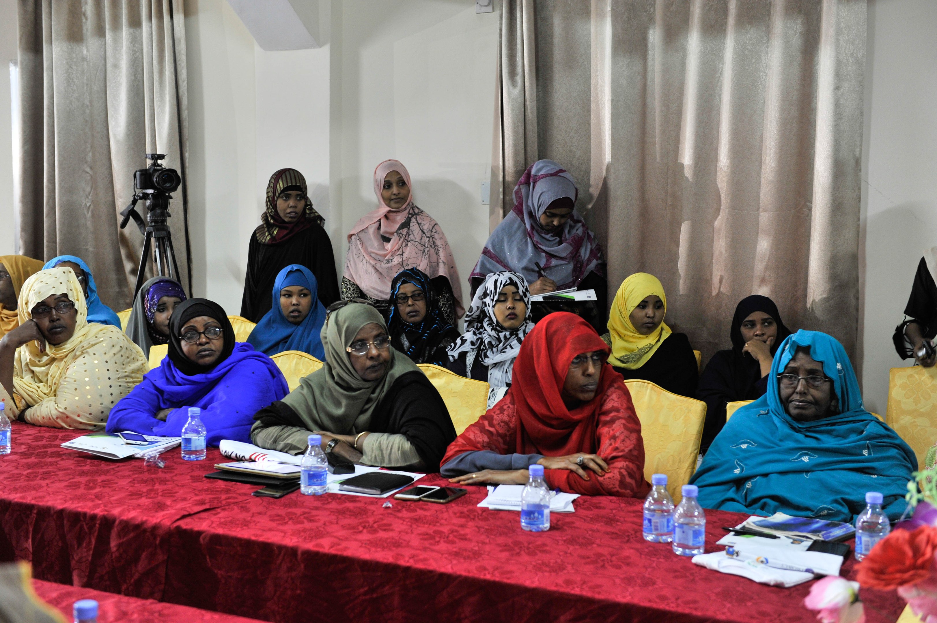 Eine Gruppe von Frauen sitzt um einen Tisch mit verschiedenen Gegenständen, die in eine Diskussion vertieft sind, mit einer Blumenvase auf der rechten Seite und Vorhängen im Hintergrund.
