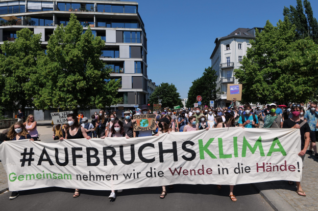 Eine Gruppe von Menschen mit Masken hält ein Banner mit der Aufschrift "Aufbruchsklima" vor einem Gebäude mit Bäumen und einem klaren Himmel, während sie gegen eine Klimapolitik protestieren.