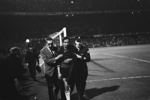 Eine Gruppe von Männern, die auf einem Fußballfeld mit einem Tor und beleuchteten Rängen im Hintergrund feiern, dargestellt in Schwarz-Weiß.
