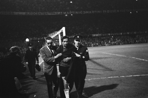 Eine Gruppe von Männern, die auf einem Fußballfeld mit einem Tor und beleuchteten Rängen im Hintergrund feiern, dargestellt in Schwarz-Weiß.