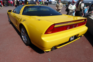 Ein gelber Sportwagen vor einer Menschenmenge geparkt, einige tragen Helme, eine Wand rechts, Zelte, Bäume, Gebäude und einen klaren blauen Himmel im Hintergrund.