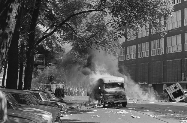 Schwarz-weiß-Foto eines brennenden Busses auf einer Straße mit parkenden Autos, Menschen, Bäumen, Gebäuden und einer Werbetafel im Hintergrund.