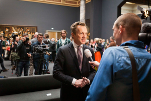 Ein Mann im Anzug und Krawatte spricht mit einem Reporter, der ein Mikrofon hält, vor einer Menge von Menschen, einige halten Kameras, ein Sofa ist im Vordergrund und eine Wand mit einem Rahmen, Lampen und einer Säule im Hintergrund.