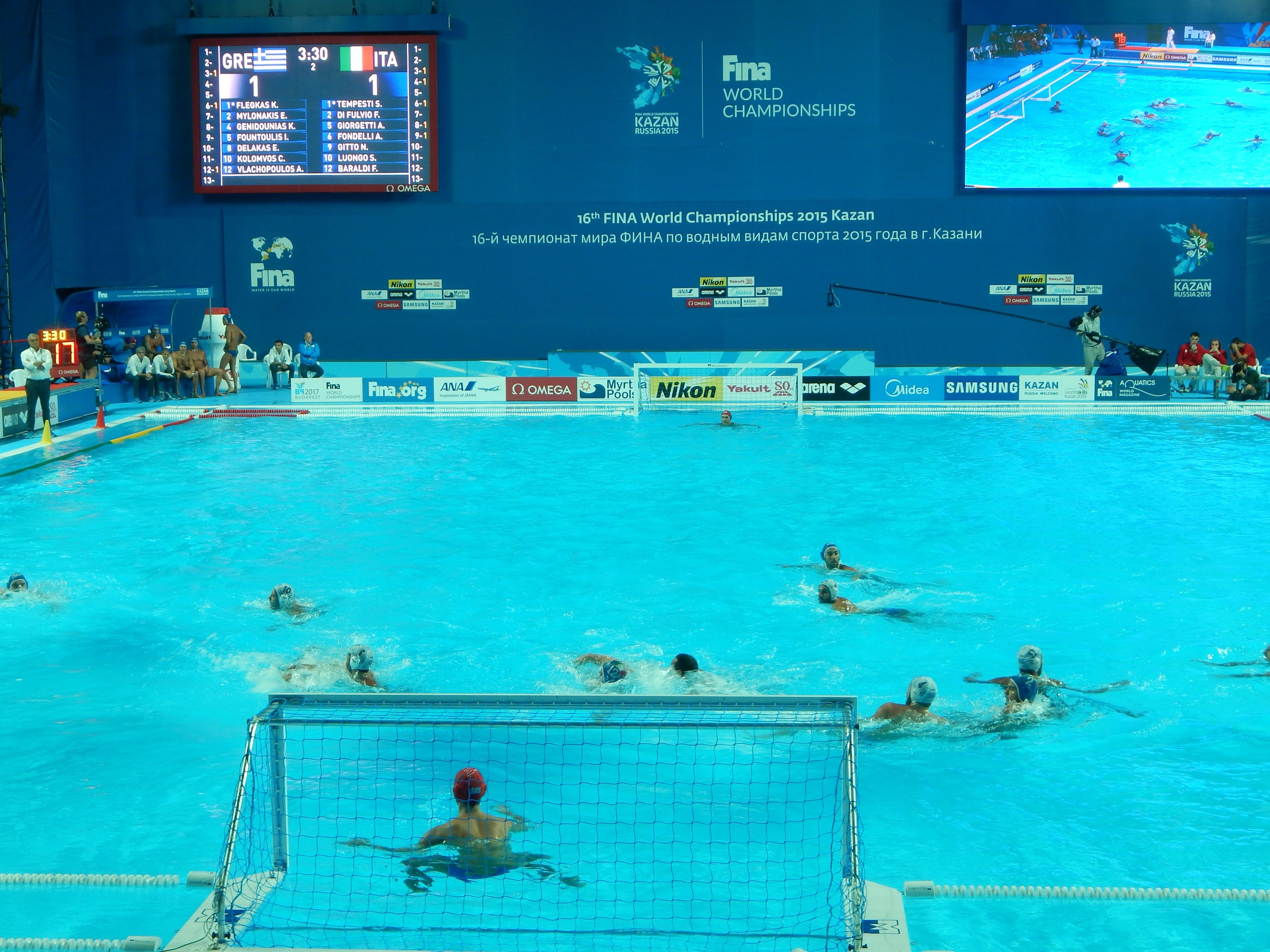 Gruppe von Athleten, die Wasserball in einem Pool während der FINA-Weltmeisterschaften 2016 in Kazan spielen, mit Zuschauern auf der linken Seite und einem Bildschirm im Hintergrund mit Event-Branding.