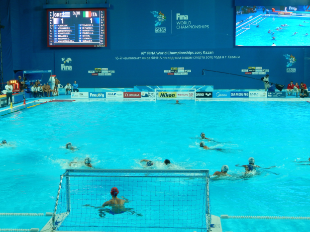 Gruppe von Athleten, die Wasserball in einem Pool während der FINA-Weltmeisterschaften 2016 in Kazan spielen, mit Zuschauern auf der linken Seite und einem Bildschirm im Hintergrund mit Event-Branding.