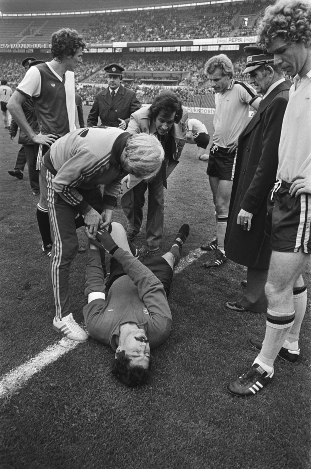 Schwarzes und weißes Bild eines Fußballspielers, der auf dem Boden liegt und von einer Gruppe Männer und einer Stadionmenge umgeben ist, wobei einer der Männer verletzt zu sein scheint.