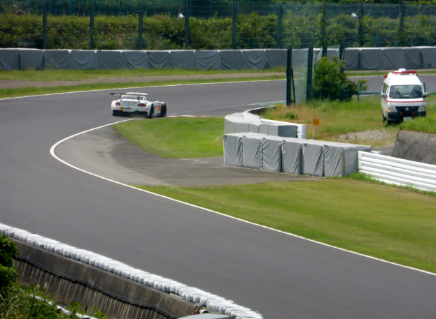 Ein Rennauto fährt eine Rennstrecke hinunter mit einem Polizeiwagen im Hintergrund, umgeben von Gras, Pflanzen, Bäumen und Zaun, mit einem Fahrzeug auf der rechten Seite und Text unten.