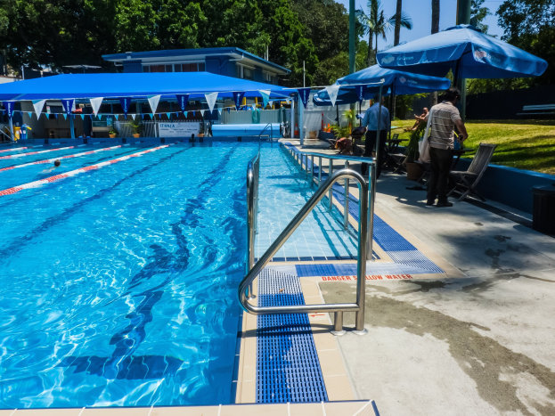 Großer Swimmingpool mit schwimmenden Menschen, umgeben von Geländern, Sonnenschirmen, Stühlen, Tischen, Pflanzen, Gras, Bäumen und einem Gebäude, unter einem klaren blauen Himmel.