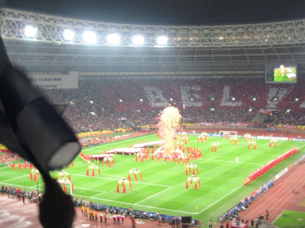 Ein Stadion mit Flutlicht, Eisengittern, Zuschauern, stehenden Personen, Werbetafeln, Bildschirmen, Überwachungskamera, Fahnen und dekorativen Elementen.