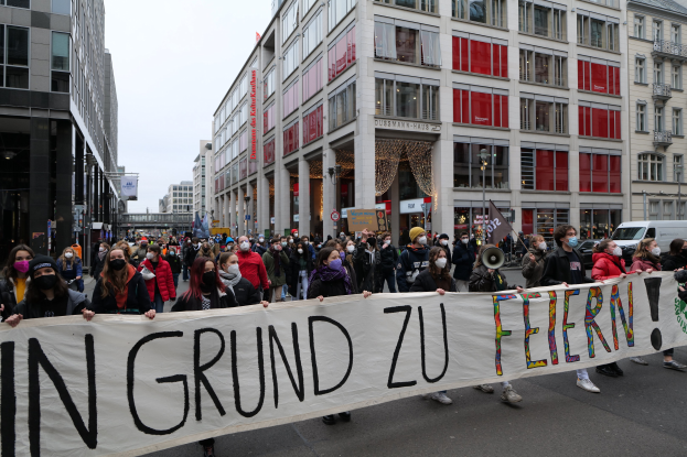Eine Gruppe von Menschen mit Masken, die ein Transparent mit der Aufschrift "In Grund zu Feiern" tragen, bei einer Demonstration in Berlin, Deutschland, mit Gebäuden, Fahrzeugen und Laternen im Hintergrund.