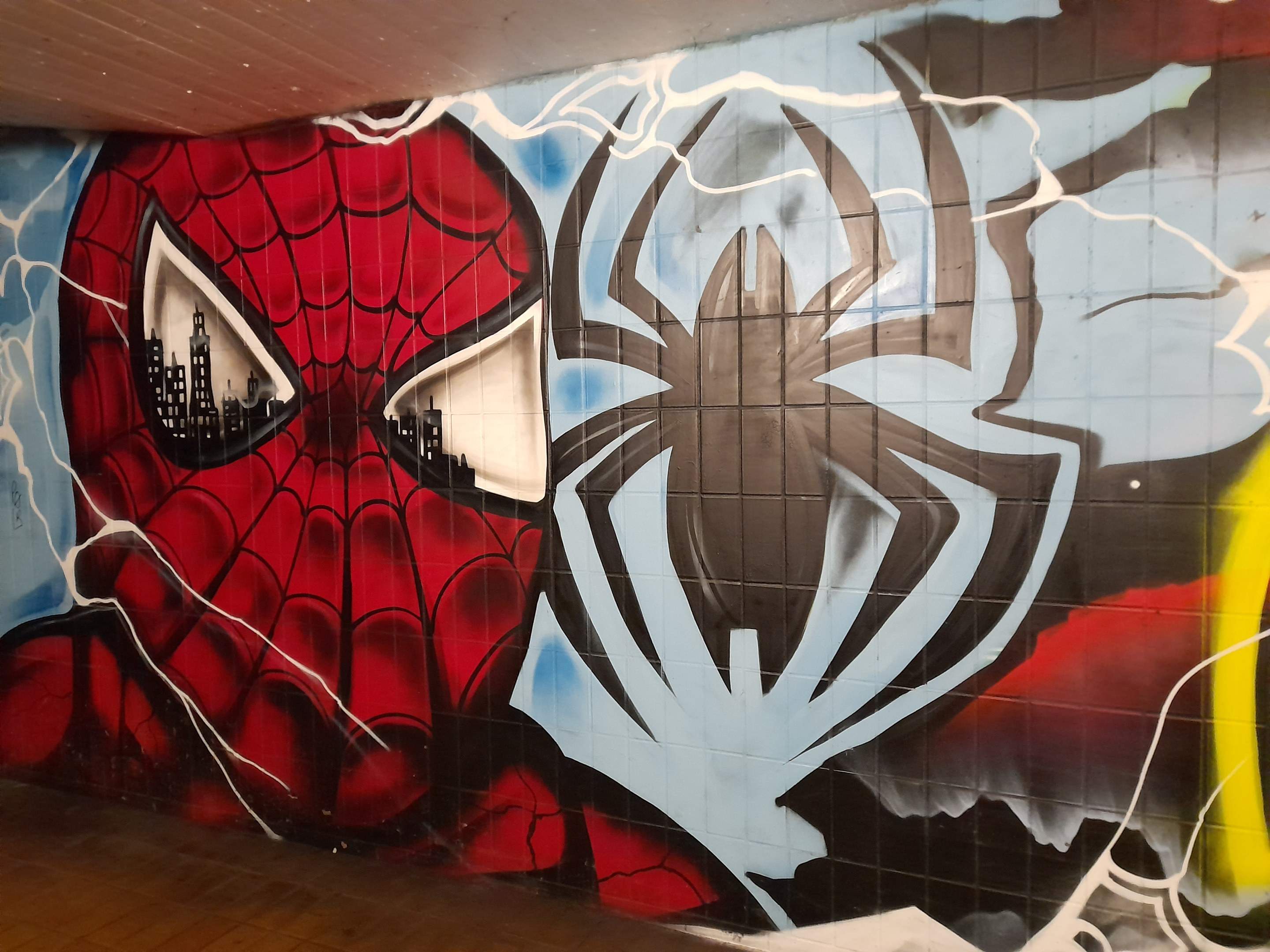 Leuchtend buntes Wandgemälde von Spider-Man in rot und blauem Kostüm auf einer Gebäudewand, mit Webmuster und großem Spinnenlogo, das ihn mit weit aufgerissenen Augen und leicht geöffnetem Mund zeigt.