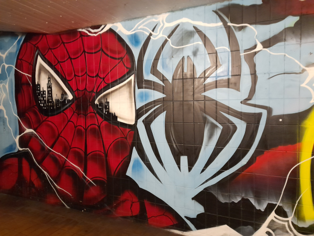 Leuchtend buntes Wandgemälde von Spider-Man in rot und blauem Kostüm auf einer Gebäudewand, mit Webmuster und großem Spinnenlogo, das ihn mit weit aufgerissenen Augen und leicht geöffnetem Mund zeigt.