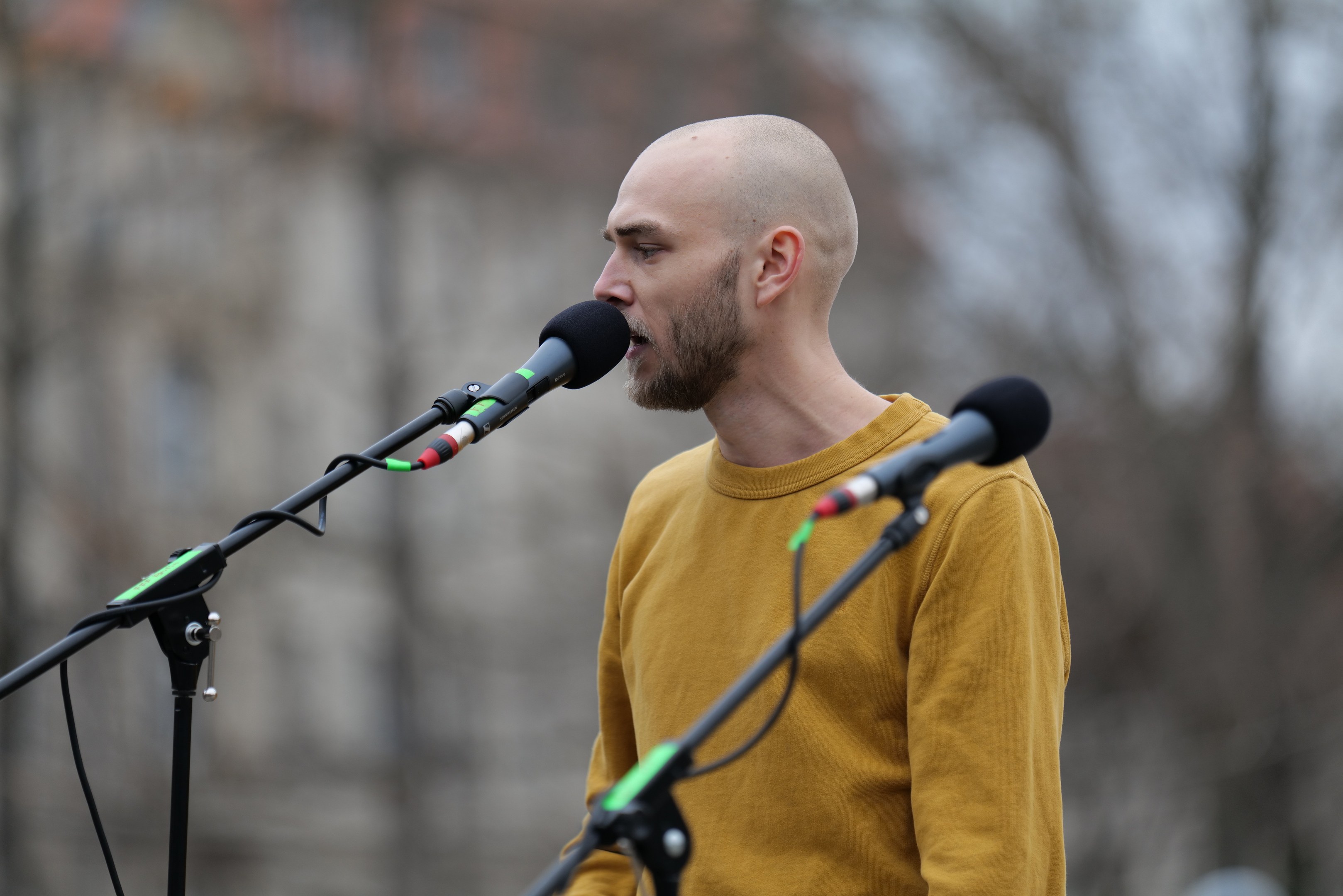 Ein kahlköpfiger Mann in einem gelben T-Shirt, der in ein Mikrofon singt, vor einem unscharfen Hintergrund.