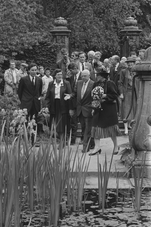 Schwarzes und weißes Bild einer Gruppe von Menschen, die sich um einen Teich in einem Garten versammelt haben, mit einer Frau in der Mitte, die einen Blumenstrauß hält, und einem Mann in einem Anzug rechts von ihr.
