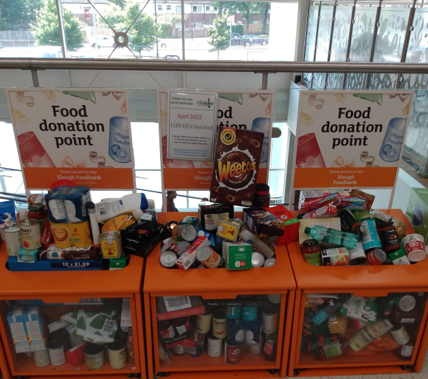 Orange Schübe mit Lebensmittelspenden gefüllt, darunter Flaschen, Dosen und Kisten, mit Informationsschildern und einem Blick auf Fahrzeuge, Bäume, Gebäude und Himmel durch Glasfenster.