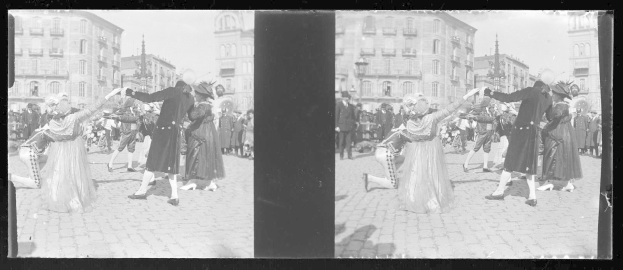 Schwarz-weiß-Foto von Menschen in Kostümen, die auf einer Straßenparade tanzen, umgeben von Gebäuden, Straßenlaternen und Lichtern unter einem klaren Himmel.