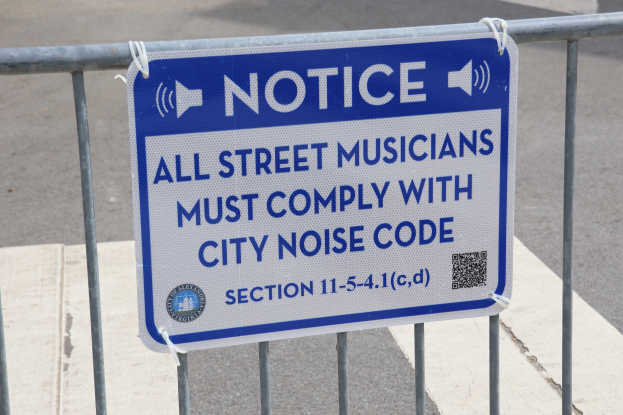 Anschlagtafel mit einem Schild, auf dem "Hinweis: Alle Straßenmusiker müssen die Stadtlärmvorschrift einhalten" steht, an einem Zaun befestigt, mit einer Straße im Hintergrund.
