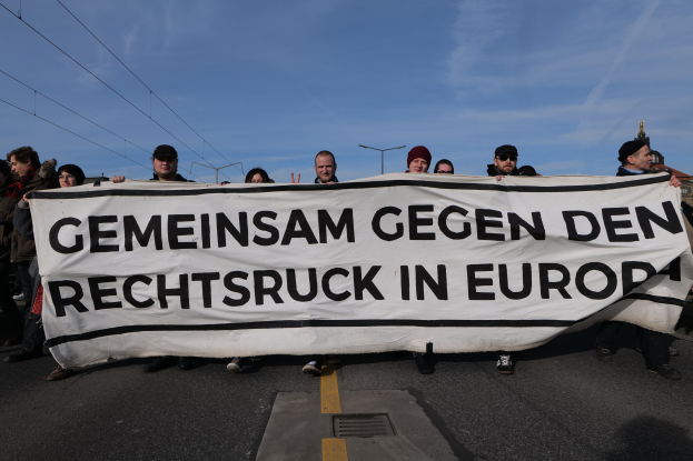 Eine Gruppe von Menschen steht auf der Straße und hält ein Banner mit der Aufschrift "Gemeinsam Gegen den Rechtsruck in Europa" hoch, mit Straßenlaternen, Strommasten, Stromkabeln, Gebäuden und einem bewölkten Himmel im Hintergrund.