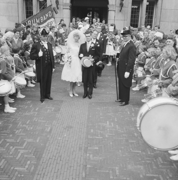 Schwarz-weiß-Foto einer Braut in einem weißen Kleid und Schleier und eines Bräutigams mit einem Blumenstrauß, der eine Straße entlang geht, vorbei an einer Menge, einige halten Musikinstrumente, mit einem Banner und einem Gebäude im Hintergrund.