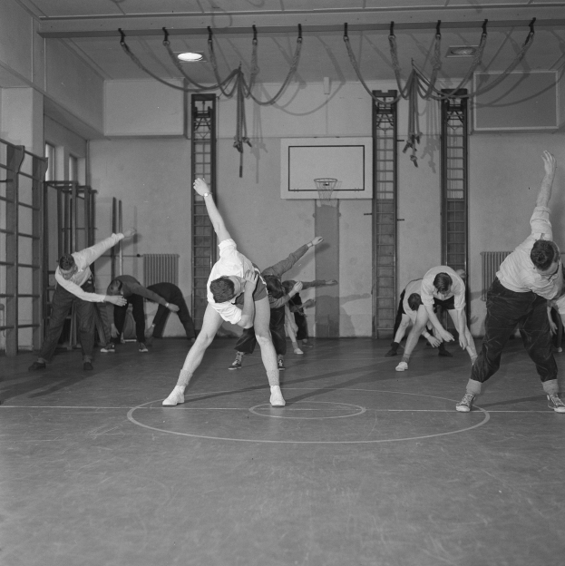 Black and white image of a group performing gymnastics in a gymnasium with a wall, ceiling lights, and floor visible.