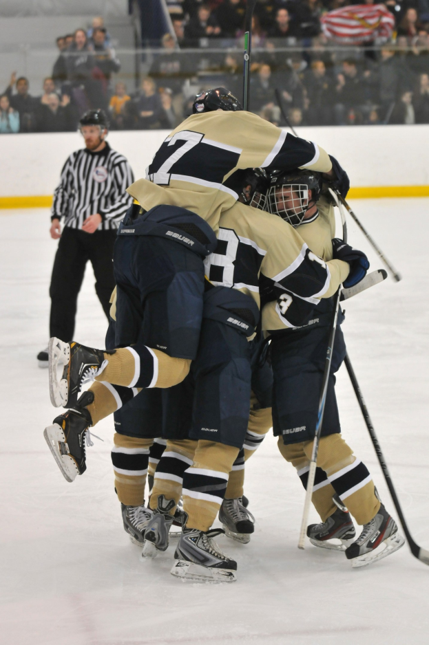 Eine Gruppe von Eishockeyspielern in Uniform und Helmen versammelt sich auf dem Eis während eines Spiels, mit Zuschauern im Hintergrund und einer Wand dahinter.
