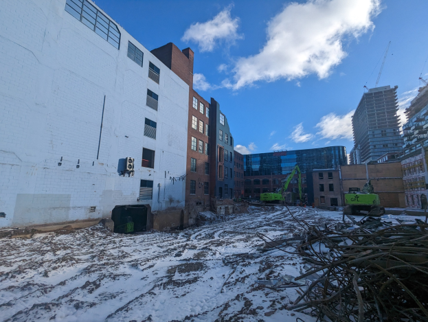 Großes Gebäude mit schwerer Schneedecke davor, umgeben von Fahrzeugen, Zweigen und städtischem Schutt, mit bewölktem Himmel und benachbarten Gebäuden im Hintergrund.
