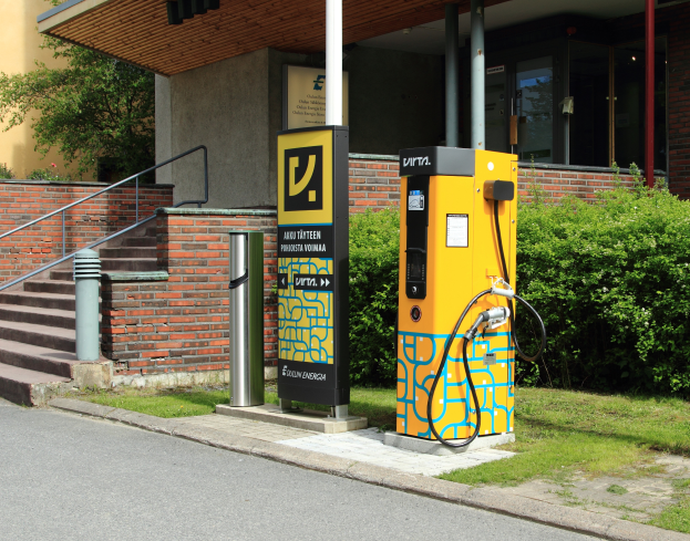 Eine Elektrofahrzeug-Ladestation an der Straße, umgeben von Grünflächen und einem Gebäude mit Fenstern und Säulen, mit Treppen und Geländern, die hinaufführen.