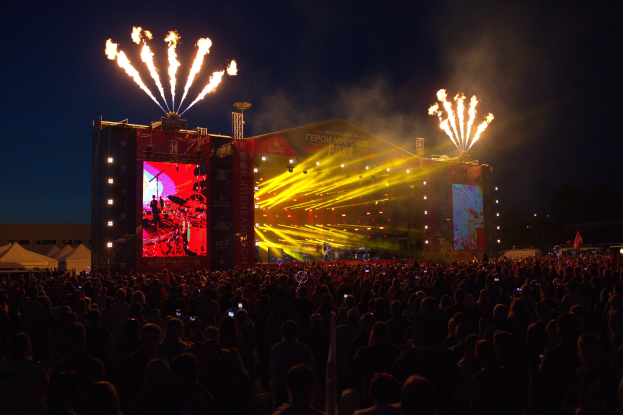 Eine große Menge bei einem Musikfestival schaut auf einer hell erleuchteten Bühne Performern zu, mit Zelten links und Feuerwerk am dunklen Himmel dahinter.